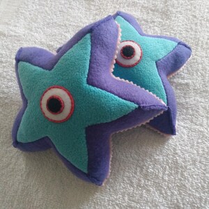 Puede incluir: Dos objetos de peluche en forma de estrella hechos de fieltro morado y verde azulado. Cada estrella presenta un diseño de ojo circular central con fondo blanco, un borde cosido en rojo y una pupila negra. Los bordes tienen costuras visibles.