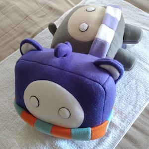 May include: Two plush toys, one purple and one gray, rest on a white towel. The purple toy is cube-shaped with a white face, blue ears, and an orange and blue striped scarf. The gray toy has a white face and a purple and white striped scarf.