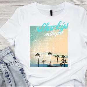 May include: White t-shirt with a graphic print of palm trees and a pier on a beach. The text "Sharky's on the pier" is printed in turquoise on the graphic.