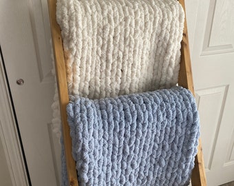 Puede incluir: Una estantería de madera en forma de escalera con dos mantas de punto. La manta superior es blanca y la manta inferior es azul claro.