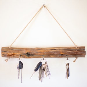 May include: A rustic wooden key holder with three metal hooks hanging from a piece of twine. The key holder is mounted on a white wall and has several keys hanging from the hooks.