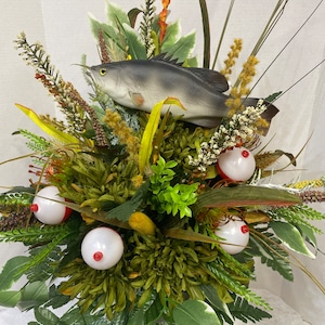 May include: A green and brown floral arrangement with a gray fish and red and white bobbers. The arrangement is in a clear vase.