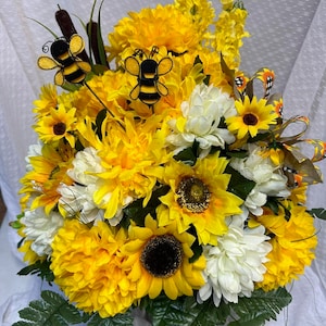 May include: A vibrant floral arrangement featuring sunflowers, yellow chrysanthemums, and white flowers. Decorative bee accents are placed throughout the bouquet. Green fern and cattail accents complete the design. A colorful ribbon adds a decorative touch.