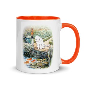 Puede incluir: Taza de cerámica blanca con borde naranja. La taza presenta una ilustración colorida de un gato blanco, un conejo marrón y una regadera en un jardín. La ilustración incluye un estanque con peces de colores.