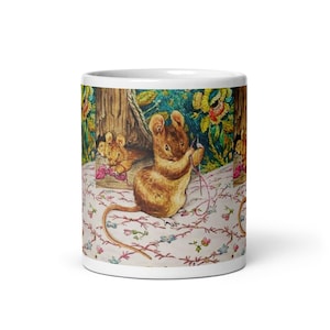 Puede incluir: Una taza de cerámica blanca con una ilustración repetida de ratones marrones en un jardín. Se muestra a un ratón cosiendo con una aguja e hilo rosa sobre una superficie de tela blanca con un patrón de pequeñas flores azules y rosas.