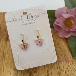 May include: A pair of gold hoop earrings with pink butterfly charms. The earrings are displayed on a card that reads "lovely things by justine earrings".