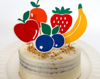May include: A white frosted cake with layers of cake and a white frosting. The cake is decorated with paper cutouts of fruit, including a red apple, an orange, a strawberry, a banana, and two blueberries.