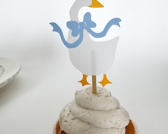 May include: A white cupcake with vanilla frosting topped with a paper goose cupcake topper. The goose has a blue bow and yellow feet.