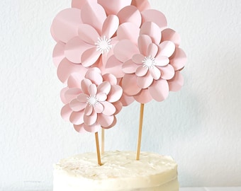 May include: A white frosted cake with pink paper flowers on top. The cake is on a gold cardboard cake base.
