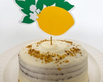 May include: A white cake with a lemon-shaped cake topper. The cake is decorated with a layer of white frosting and a sprinkle of brown crumbs. The lemon topper is yellow with green leaves and white flowers.
