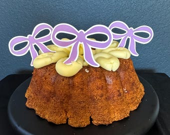 May include: A brown bundt cake with yellow frosting and purple bow decorations. The cake sits on a black plate against a dark blue background. The cake has a fluted design and is decorated with several bow-shaped toppers.