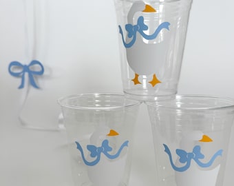 May include: Three clear plastic cups with a white goose design with a blue bow. The goose has yellow feet and a yellow beak.