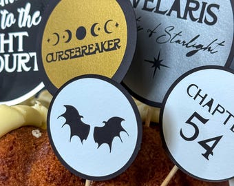 May include: Cake toppers with various designs and text, including "CURSEBREAKER", "VELARIS", and "CHAPTER 54". The toppers are round with black, gold, and light blue backgrounds. Some feature moon phases and bat silhouettes.
