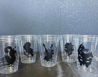May include: A set of clear plastic cups with black silhouette designs. Each cup features a different stylized figure. The cups are arranged on a dark surface against a light blue background.