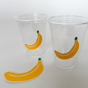 Puede incluir: Tres vasos de plástico transparentes con pegatinas de plátano amarillo. Los plátanos tienen tallos verdes.