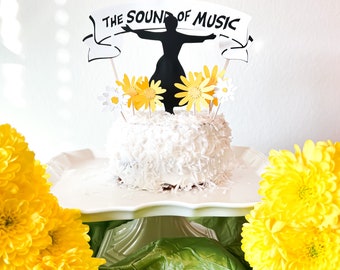 May include: A white cake with coconut flakes on top, decorated with a black silhouette of a woman dancing and a banner that says "The Sound of Music". The cake is surrounded by yellow flowers.