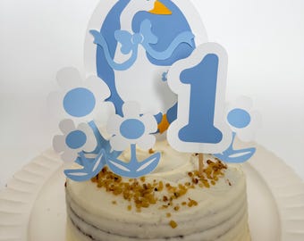 May include: A white cake with blue and white decorations. The cake is decorated with a blue and white goose, flowers, and the number "1".