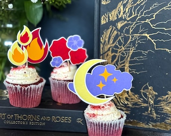 May include: Three red velvet cupcakes with white frosting and decorative toppers. The toppers include flames, red roses, purple flowers, a yellow crescent moon, and stars. The cupcakes are arranged in front of a book with gold lettering.