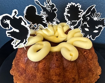 May include: A bundt cake with light brown sides and a yellow frosting design. The cake is decorated with black and white silhouette cutouts of various figures. The cake sits on a black plate against a blue background.