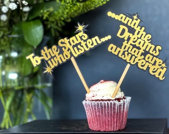 May include: A red velvet cupcake with white frosting is decorated with two gold glitter toppers that read "To the Stars Who Listen..." and "...and the Dreams that are answered." The cupcake sits on a book titled "A Court of Thorns and Roses."