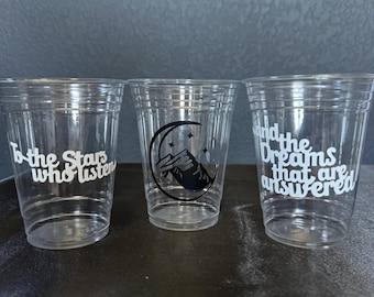 May include: Three clear plastic cups with white text and black graphics. One cup reads "To the Stars who listen", another features a mountain and moon design, and the third says "and the Dreams that are answered".