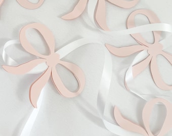 May include: A garland of five pink paper bows tied with white ribbon. The bows are cut out in a simple shape and are strung together with white ribbon.