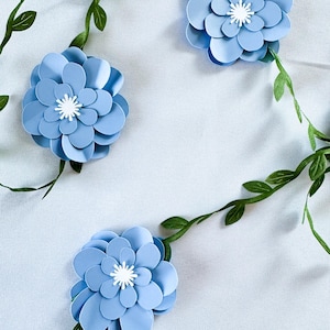 Può includere: Tre fiori di carta azzurri con centri bianchi sono disposti su una superficie bianca, con viti verdi e frondose intrecciate. I fiori hanno più strati di petali, creando un effetto dimensionale. Il tema generale è floreale e decorativo.