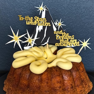 May include: A bundt cake with yellow frosting and a decorative topper. The topper features a black mountain silhouette, gold stars, and the text "To the Stars who listen and the Dreams that are answered."