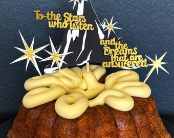 May include: A bundt cake with yellow frosting and a decorative topper. The topper features a black mountain silhouette, gold stars, and the text "To the Stars who listen and the Dreams that are answered."