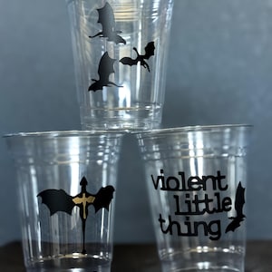 Peut inclure: Trois gobelets en plastique transparents empilés, chacun avec un motif de chauve-souris noir. Un gobelet porte l'inscription "violent little thing". Les gobelets sont sur une surface gris foncé.