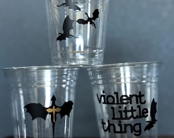 May include: Three clear plastic cups stacked, each with a black bat or bat-like design. One cup has the text "violent little thing". The cups are on a dark gray surface.