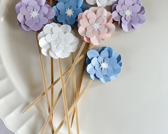 May include: A set of eight paper flower cake toppers in shades of pink, blue, and white. The flowers are attached to wooden skewers.