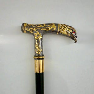 May include: A decorative walking cane with a black shaft and a detailed handle. The handle features a gold-colored eagle head with red eye accents and ornate gold filigree designs on a dark gray background.