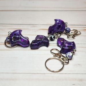 以下が含まれることがあります： 紫色の樹脂製キーホルダー4つ。星座のデザインです。各キーホルダーには、シルバーのキーリングとロブスタークラスプが付いています。キーホルダーは明るい木製の表面にあります。