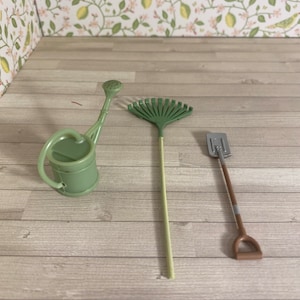 Puede incluir: Un juego de jardinería en miniatura con una regadera verde, un rastrillo y una pala. La regadera tiene un asa y un pico. El rastrillo tiene una cabeza en forma de abanico. La pala tiene un mango marrón y una hoja plateada.