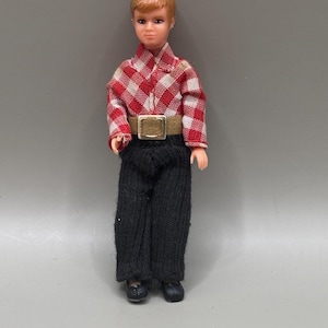 Puede incluir: Una muñeca vintage de una persona joven con pelo corto, rubio rojizo. La muñeca lleva una camisa a cuadros rojos y blancos, pantalones negros, un cinturón marrón con hebilla dorada y zapatos negros. La muñeca está sobre un fondo gris.