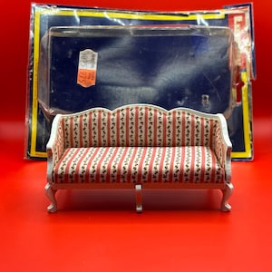 May include: Miniature sofa with a white frame and red and white striped upholstery. The sofa has curved legs and a detailed back. The sofa is in front of a blue and yellow package on a red surface.