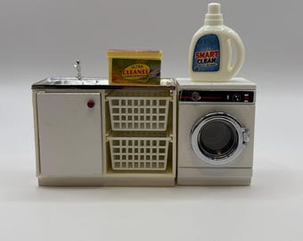 Może przedstawiać: Miniaturowy zestaw pralni z białą pralką, zlewem i szafką z dwoma koszami na pranie. Obecna jest również butelka detergentu "SMART CLEAN" i pudełko detergentu "ULTRA CLEANEL".