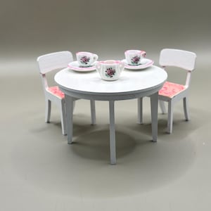 May include: A miniature white table with three matching chairs, set for tea. The table holds two teacups and saucers, plus a creamer, all decorated with pink roses. The chairs have pink floral patterned seats.