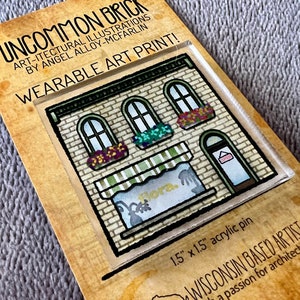 May include: A wearable art print of a brick building with a storefront. The storefront has a sign that says "Flora" and a striped awning. The building is 1.5 inches by 1.5 inches and is made of acrylic.