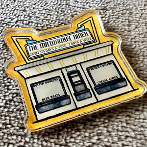 Op de afbeelding: Een gele en witte illustratie van een diner met de tekst "THE MILWAUKEE DINER" en "OPEN 365 DAYS A YEAR 7 DAYS A WEEK". Het diner heeft drie ramen met de tekst "BUTTER BURGERS", "BEER BRATS", "FROZEN CUSTARD" en "CHEESE CURDS".