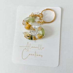 May include: A clear resin letter "S" keyring with gold-coloured hardware. The keyring is filled with dried yellow and green flowers and gold flakes. The keyring is attached to a gold ring. The card behind the keyring has the text "Almarillo Creations".