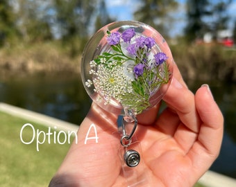 May include: A clear, round badge reel with a floral design. The reel contains dried purple and white flowers encased in resin. The text "Option A" is written in white cursive on the left side of the image.