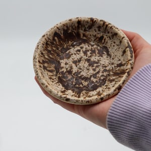 Könnte beinhalten: Eine kleine, runde Keramikschale mit einem gesprenkelten braunen und beigen Design. Die Schale wird in einer Hand gehalten und zeigt das einzigartige, handgefertigte Aussehen. Innen und außen ist ein Spritzmuster zu sehen, das für visuelles Interesse sorgt.