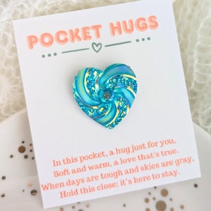 May include: A small blue heart-shaped charm with glitter and a swirl design. The charm is on a white card with the text "POCKET HUGS... - ..." and a message about a hug in your pocket.