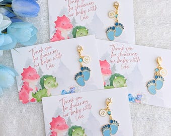 Op de afbeelding: Vier blauwe en witte babyvoet-sleutelhangers met een gouden bedel bevestigd aan een kaart met de tekst "Bedankt voor het vieren van onze baby met liefde". De kaart heeft een aquarel-illustratie van twee dinosaurussen.