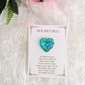 May include: A teal heart-shaped pin with gold accents, displayed on a card that reads "POCKET HUG." The card includes a poem about comfort and love. The background is a soft, white, textured fabric.
