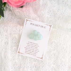 Puede incluir: Una tarjeta pequeña con el texto "Pocket Hug" y un amuleto en forma de nube de color azul claro y brillante. La tarjeta dice "En este bolsillo, un abrazo solo para ti. Suave y cálido, un amor que es verdadero. Cuando los días son difíciles y el cielo está gris, mantén esto cerca. Está aquí para quedarse."