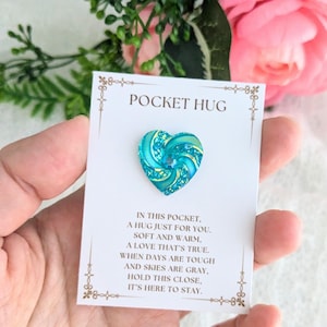 May include: A small turquoise heart-shaped pocket hug with a sparkly swirl design and the text "Pocket Hug" on a white card with a gold border. The card reads "In this pocket, a hug just for you. Soft and warm, a love that's true. When days are tough and skies are gray, hold this close, it's here to stay."