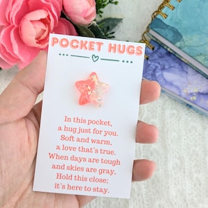 May include: A small white card with the text "POCKET HUGS" and a pink and white glittery star charm attached. The card has a poem that reads: "In this pocket, a hug just for you. Soft and warm, a love that's true. When days are tough and skies are gray, Hold this close; it's here to stay."